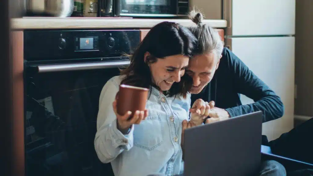 A couple sitting on the kitchen floor laughing over a laptop.