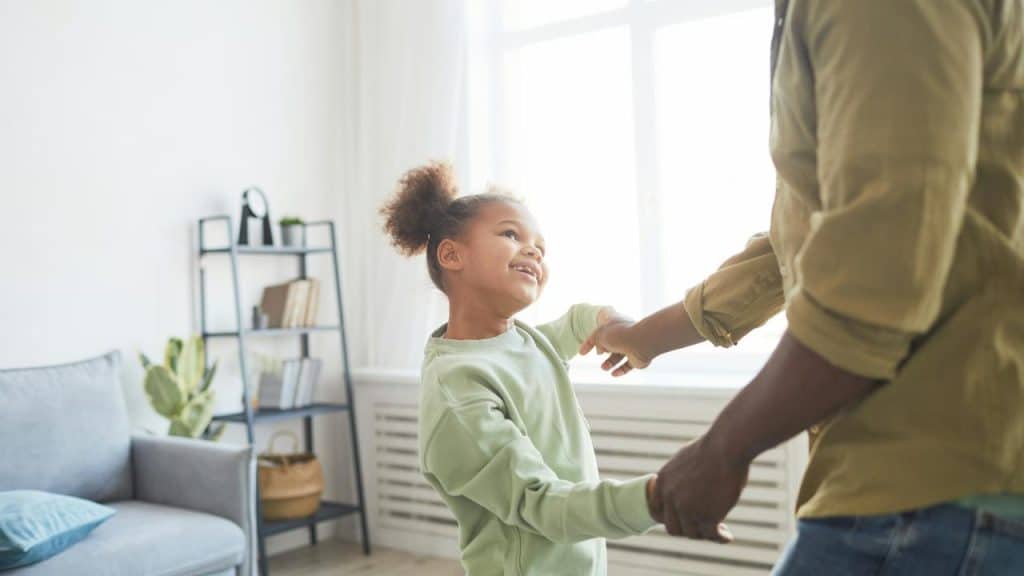 A child smiling and holding hands with an adult while dancing in a bright living room.