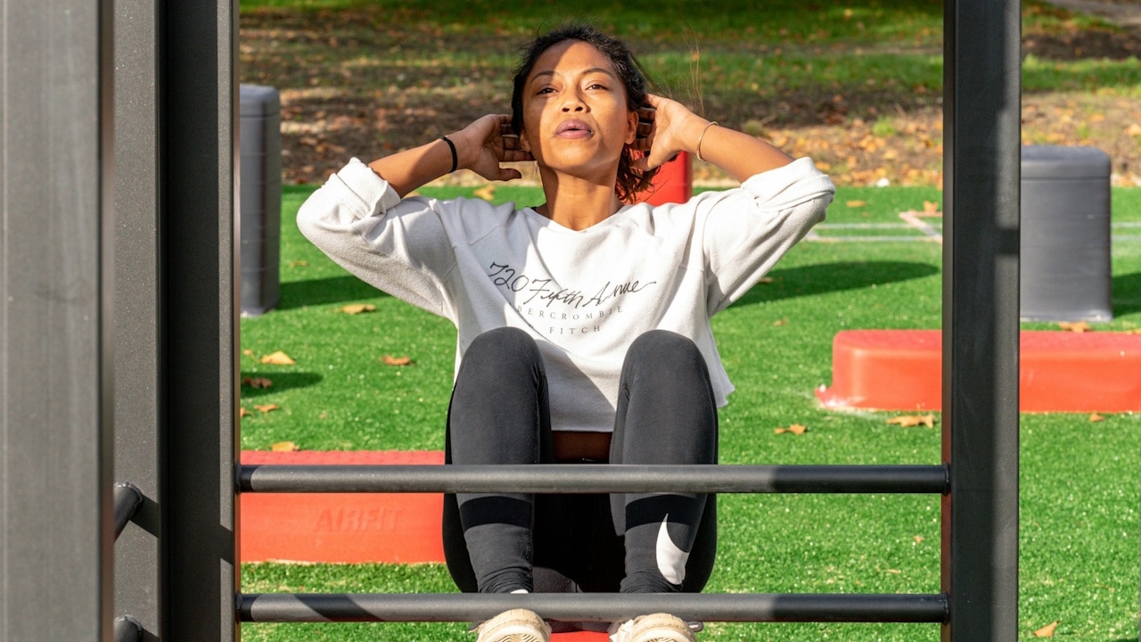 A woman is doing sit-ups on an outdoor fitness apparatus with her hands behind her head.