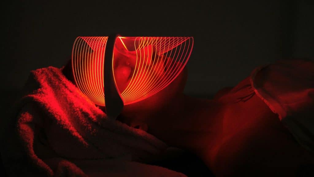 A person is lying down wearing an LED light therapy mask glowing bright red.