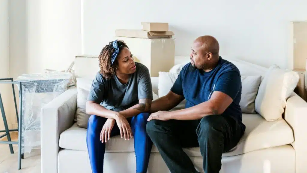 A Black couple is sitting on a white couch, talking with moving boxes behind them.