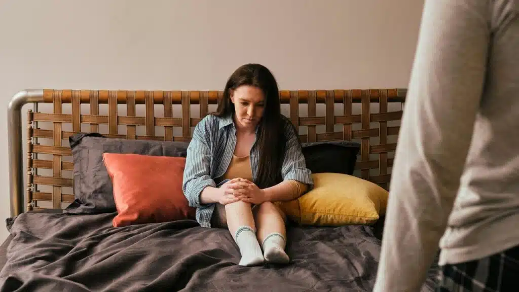 Woman sitting alone on a bed with a man's back visible in the foreground.