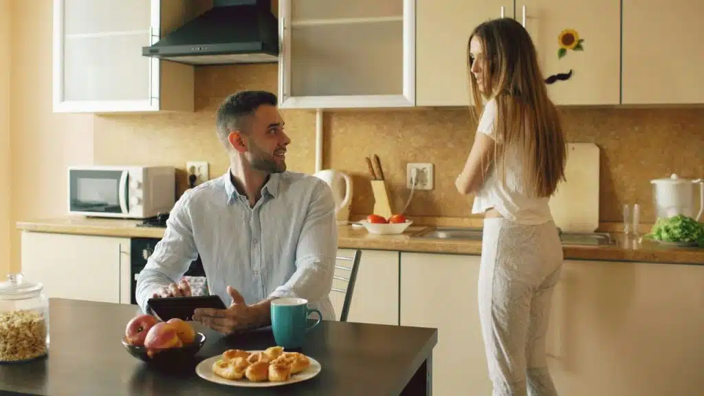 Man sitting at a table with a tablet, woman standing and talking to him in a kitchen.