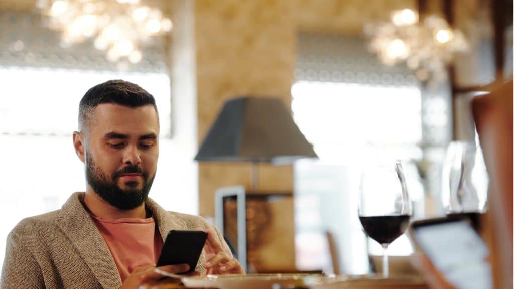 A man with a beard, wearing a blazer over a pink shirt, looks at his phone while sitting at a dining table in a restaurant.