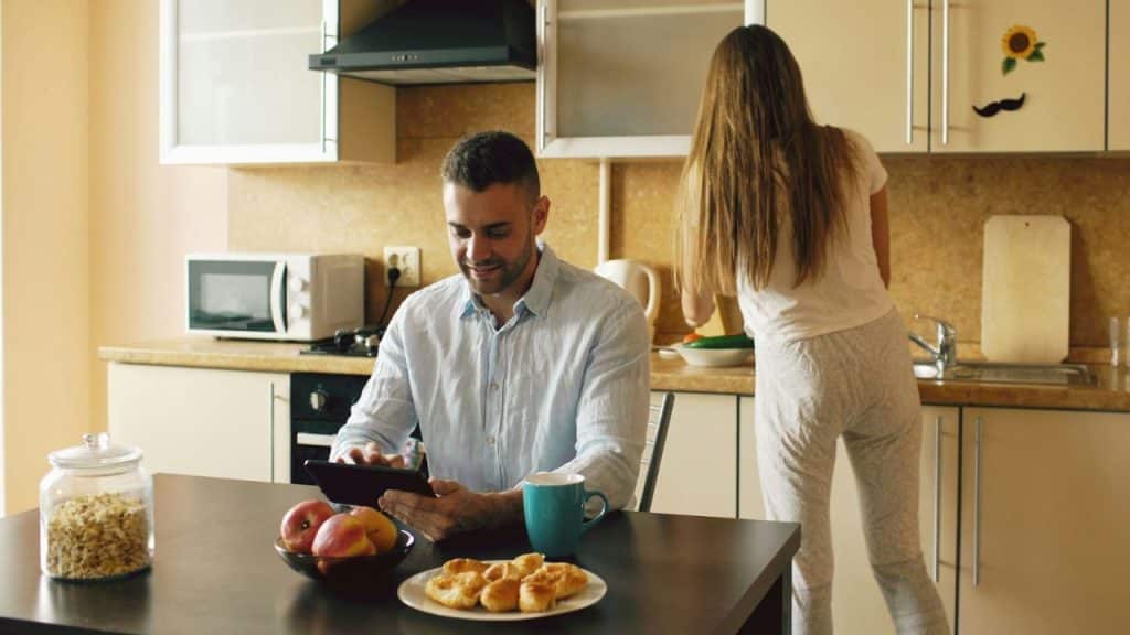 A man smiles at a tablet while a woman works with her back to the kitchen counter.