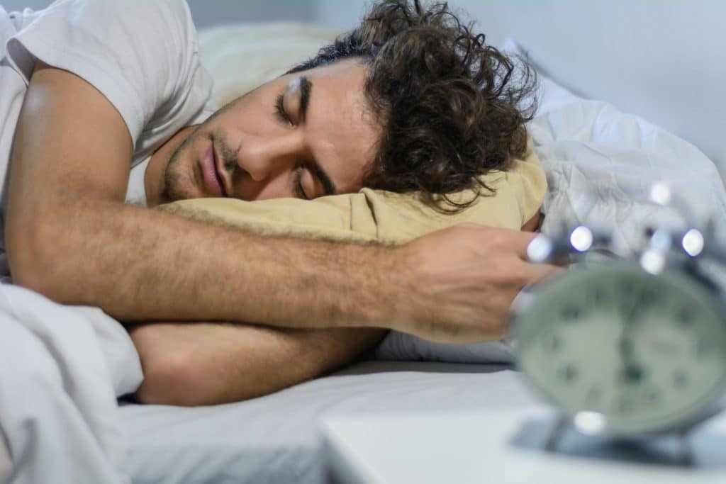 A man sleeping with an alarm clock on his bedside table.