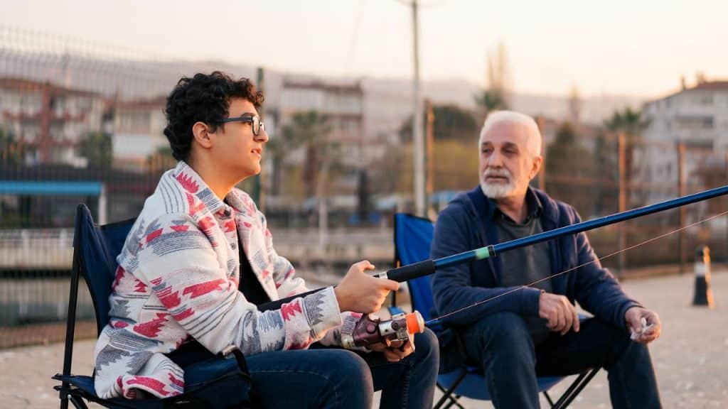 A young man and a man talking while fishing by the water in folding chairs at sunset.