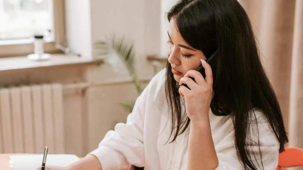 A woman talking on the phone while writing at a desk.