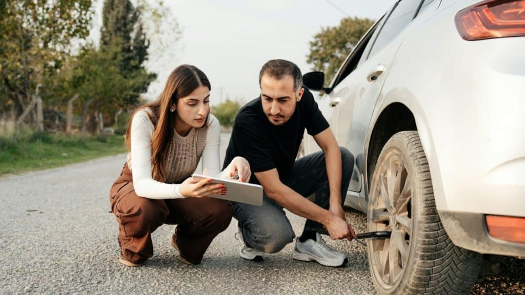 A man kneels by a car tire while a woman points to a tablet on a roadside.