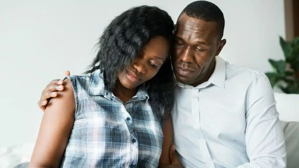 Black man comforting a black woman with his arm around her shoulder indoors.