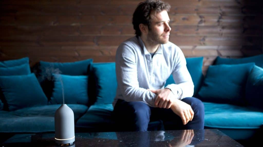 A man sits on a teal velvet couch, looking away from a smoking essential oil diffuser.