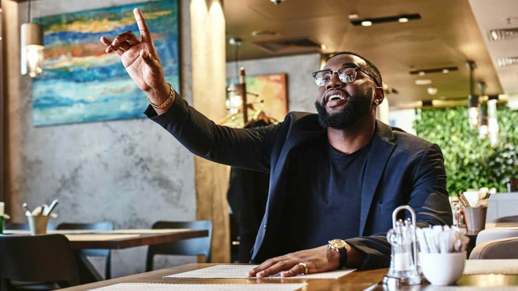 A man in a suit jacket and glasses raises his hand to signal staff in a restaurant.