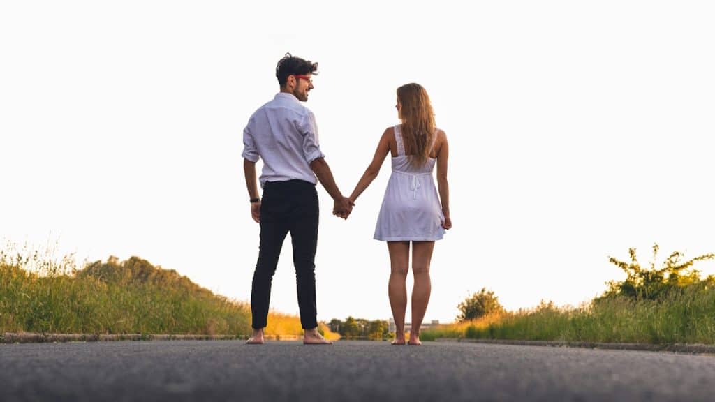 A man and a woman stand barefoot on a paved road, holding hands and looking toward a bright, sunlit horizon bordered by tall grass.