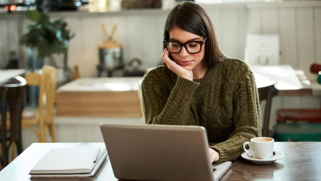 Woman in an olive green sweater and glasses working on a laptop in a cafe.