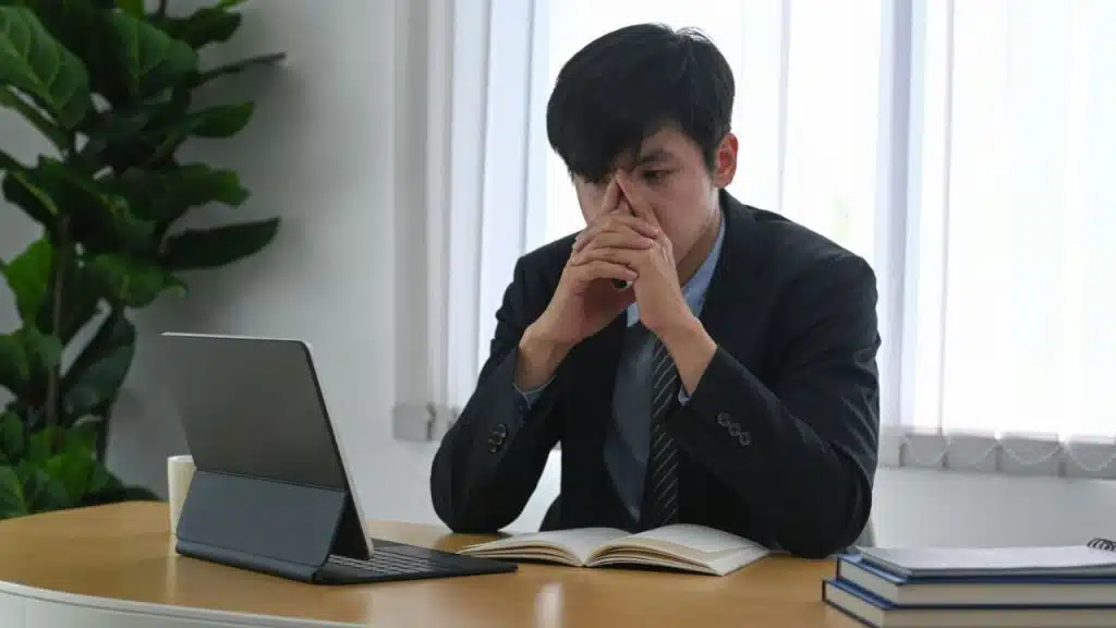 Asian man in a suit jacket sits at a desk, looking troubled with hands on face.