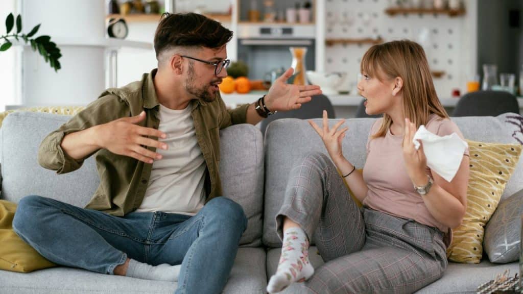 Man and woman arguing on a couch, both gesturing intensely with their hands.
