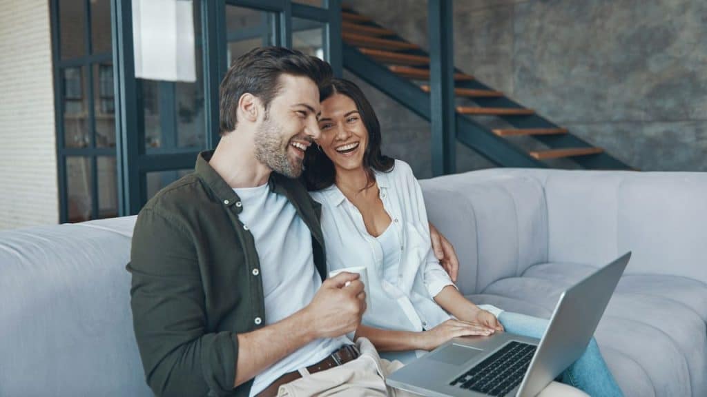 Smiling couple on a gray couch using a laptop together in a modern, well-lit living room.