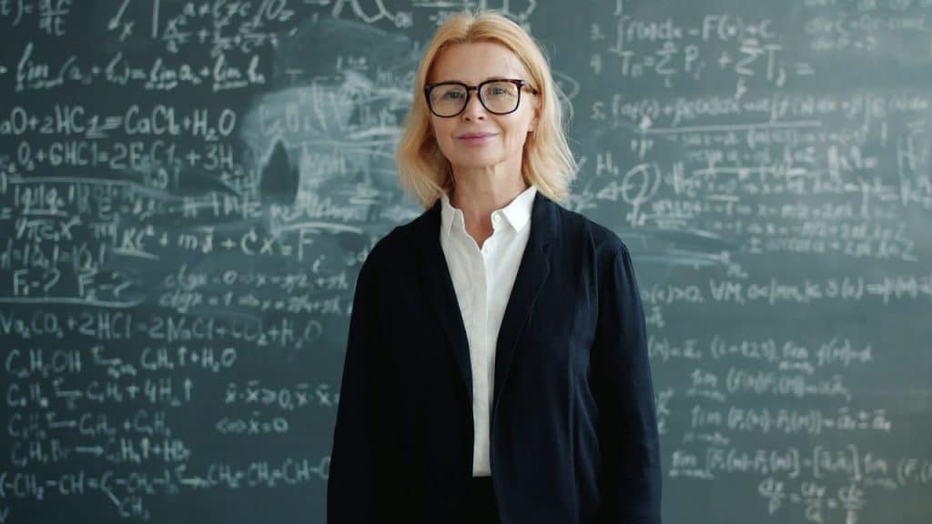 A confident woman wearing glasses stands in front of a chalkboard full of equations.