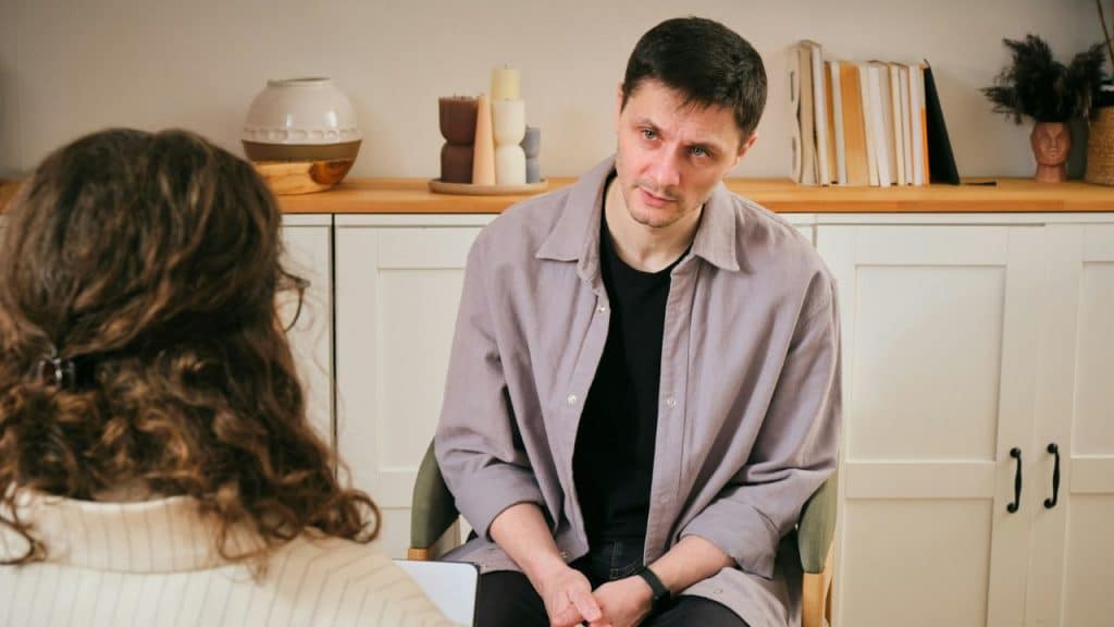 A man in a gray shirt sits attentively listening to a person opposite him.