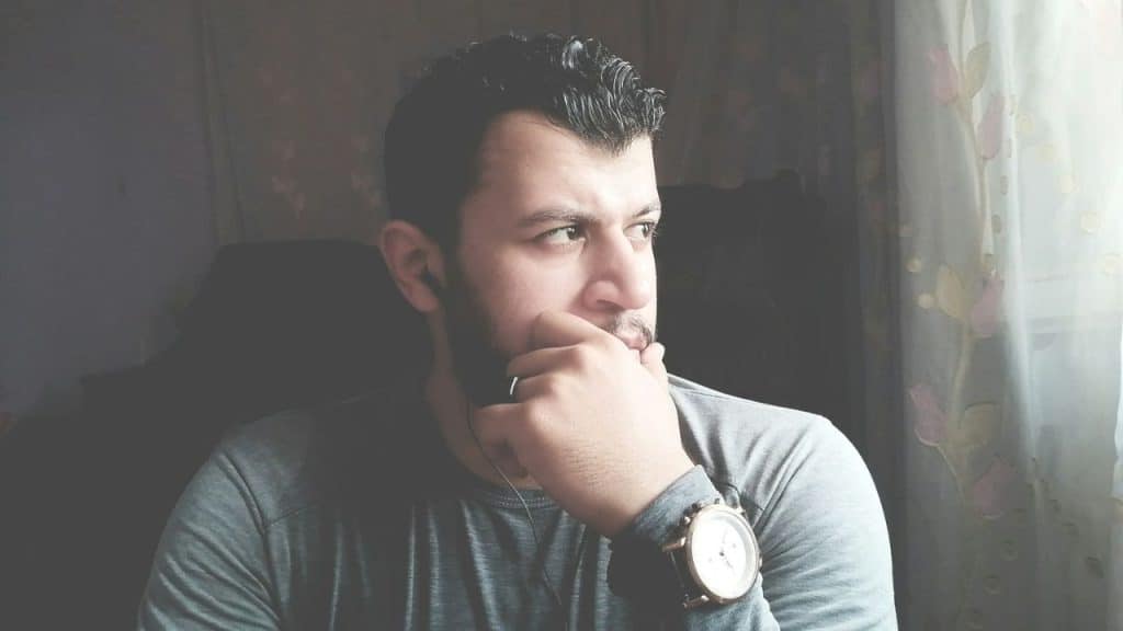 A man with a beard, wearing a gray shirt and a watch, is looking out a window.