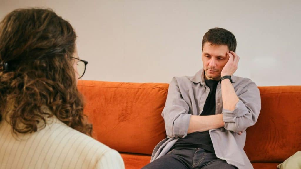A man sits on an orange couch, looking down while speaking to a woman.