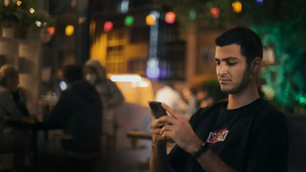 A young man sits outdoors at night, looking at his phone with colorful blurred lights in the background.