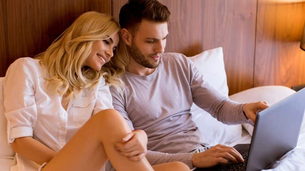 A blonde woman and a man in bed looking at a laptop computer together.