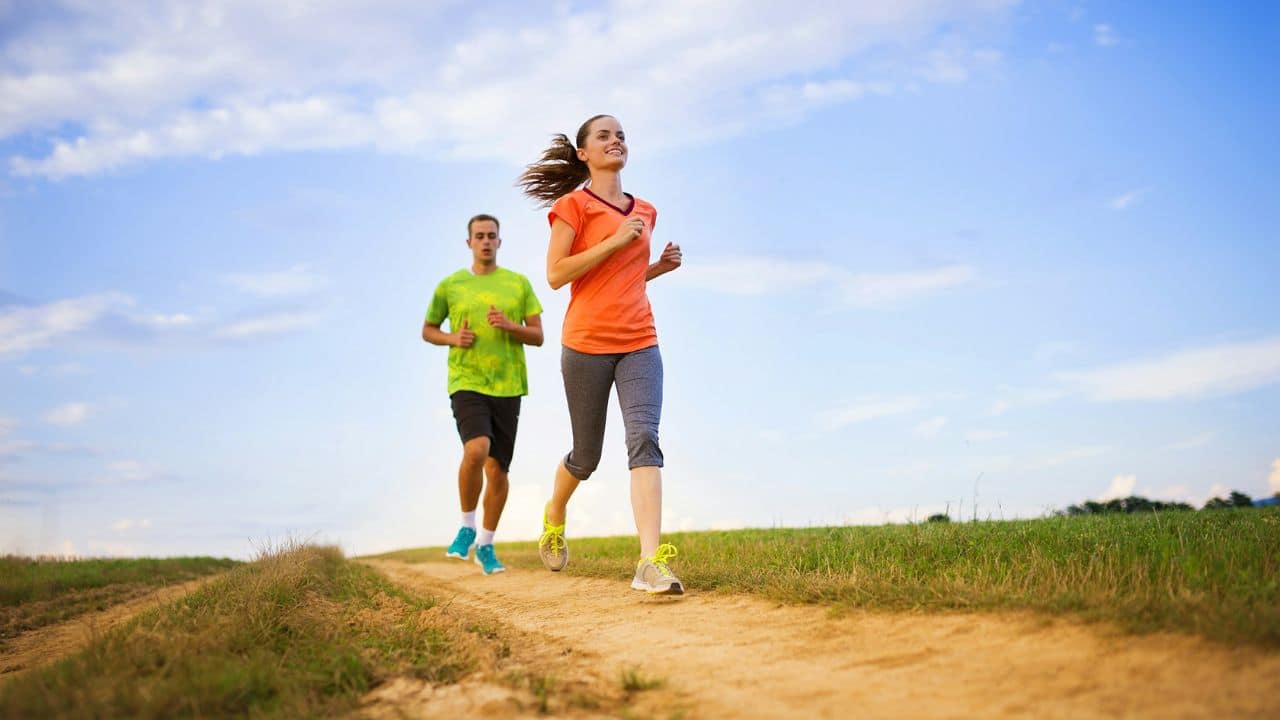 A man and a woman jogging on a dirt trail under a bright blue sky.