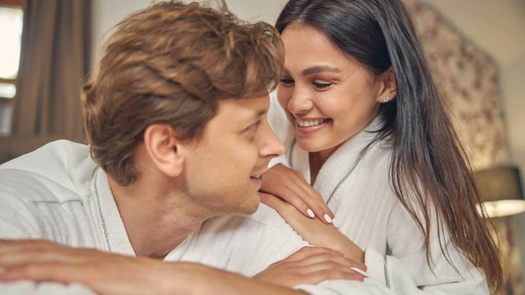Smiling couple in white bathrobes looking affectionately at each other in bed.