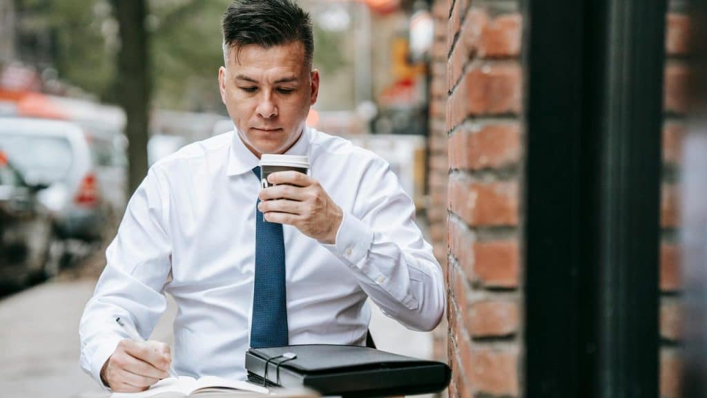 A man drinking coffee while writing at an outdoor table.