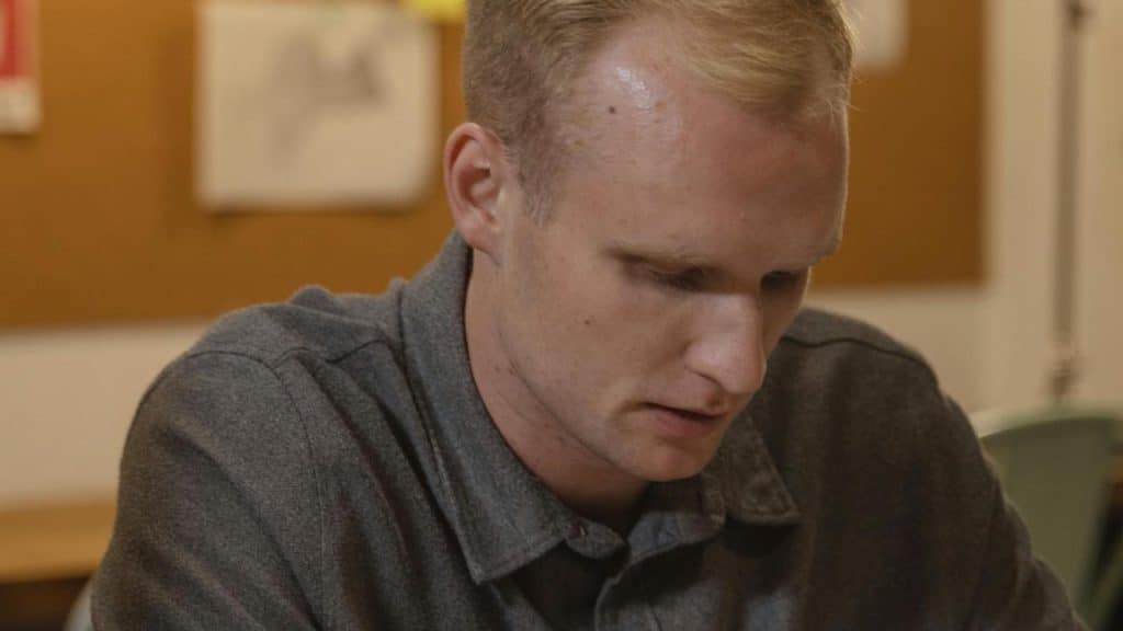 A man with short blond hair looking down while sitting indoors.