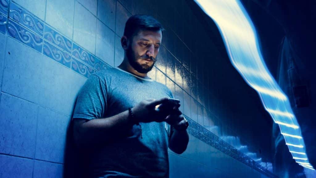 Man with beard and mustache in a blue-lit area checking his phone against a tiled wall.