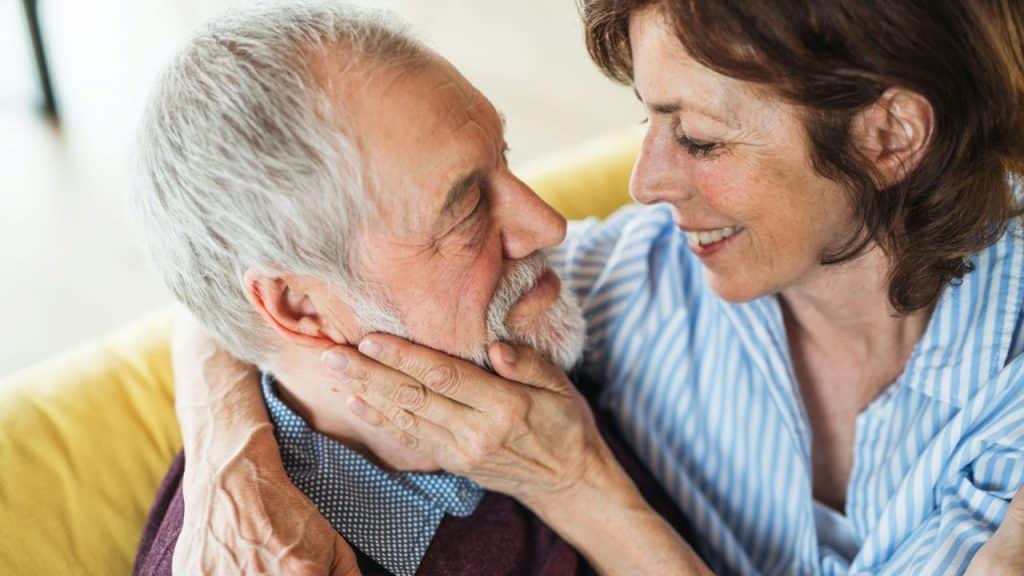 Elderly woman gently holds a man's face while looking at him and smiling.