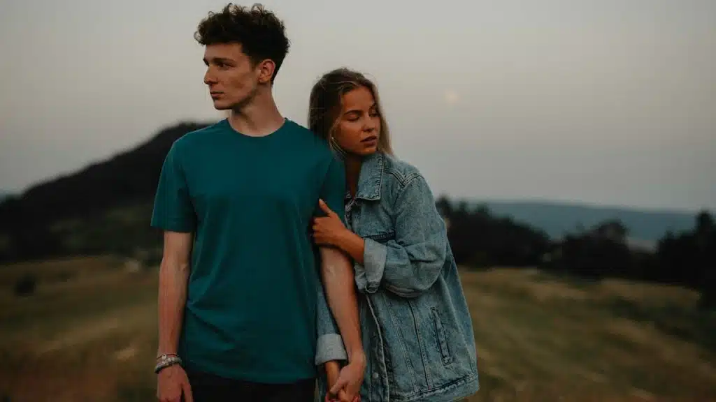 A young couple standing in nature in the countryside, holding hands but looking away from each other.