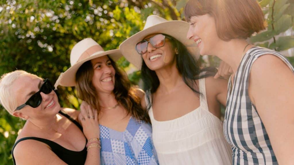 Four women in sun hats and summer clothes are laughing together outdoors in the sun.