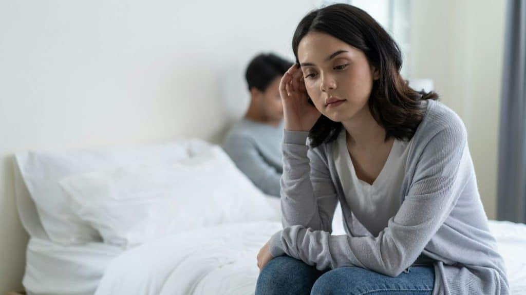 Woman in grey cardigan sits on bed, looking down, man sits behind her, blurred.