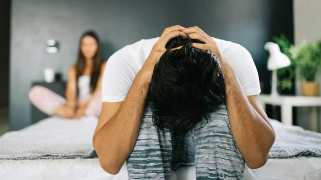 A distressed man sits on a bed with his head in his hands; a blurred woman sits in the background.
