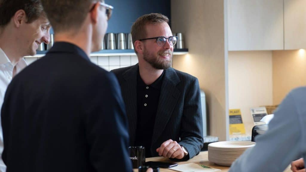 Man with a beard and glasses smiling while talking to other men indoors.