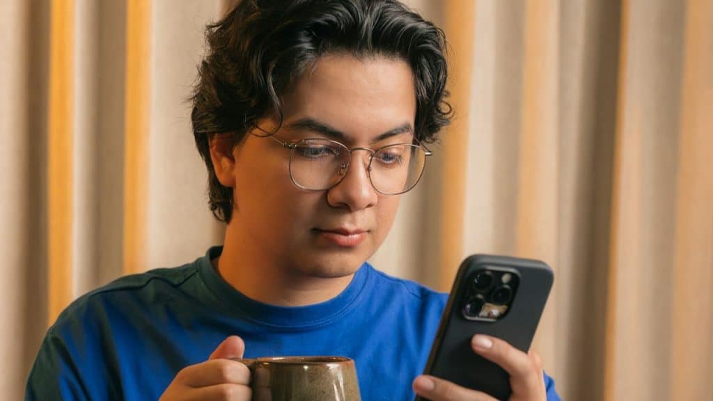 Young man in glasses and blue shirt looking at his smartphone while holding a mug.