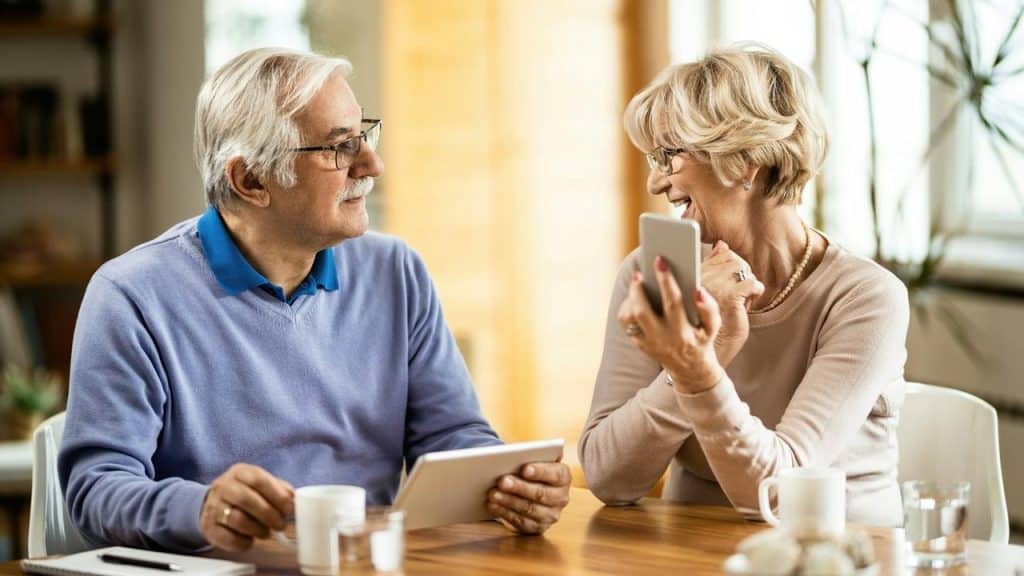 An older couple smiling and chatting while using a tablet and smartphone.