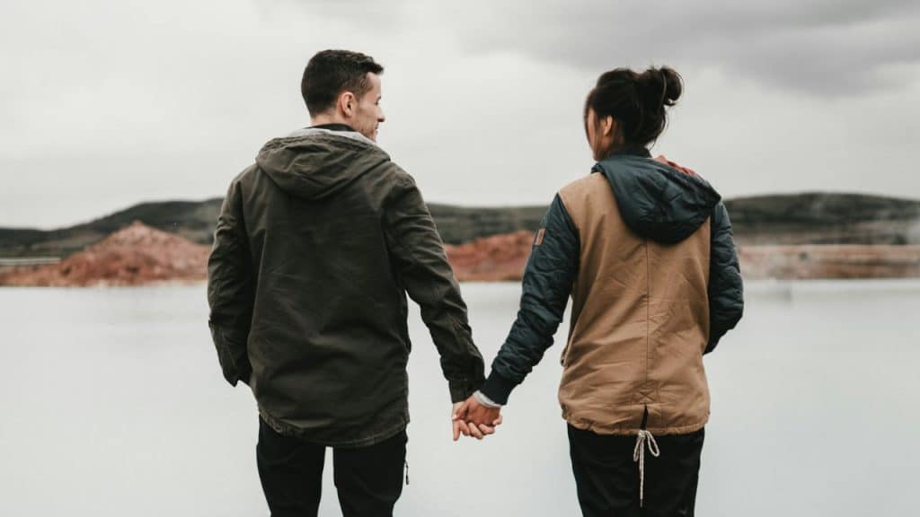 A man and woman hold hands while standing outdoors looking at a body of water.
