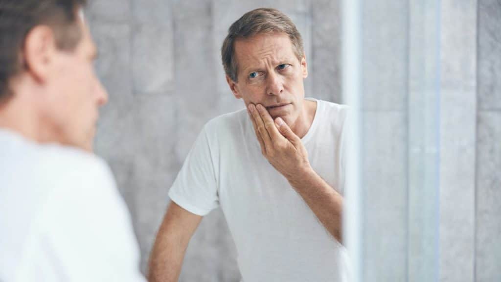 A middle-aged man in a white t-shirt touches his face while looking in a mirror.