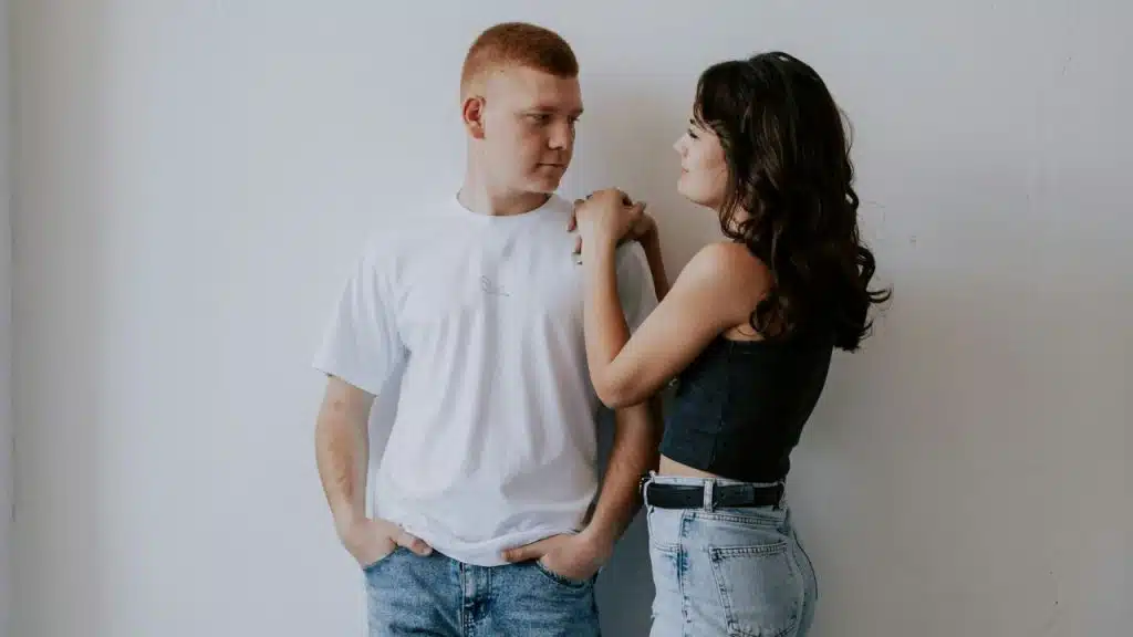 A woman in a dark crop top rests her hand on a man's shoulder against a white wall.