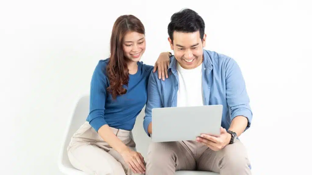 A man and woman sit side-by-side on a white chair while smiling and looking at a laptop.