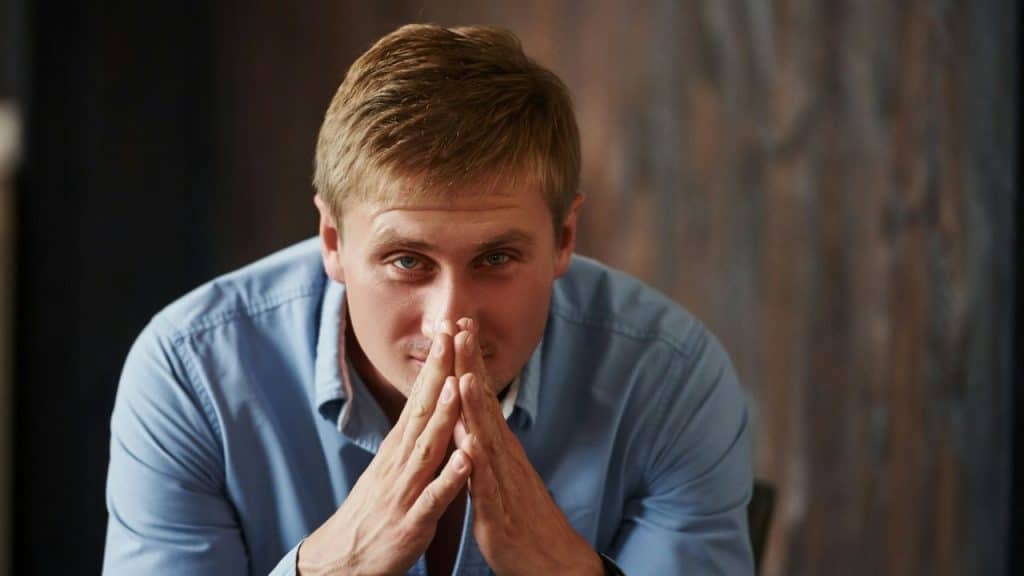 A man sits with his hands clasped together, looking directly at the camera.