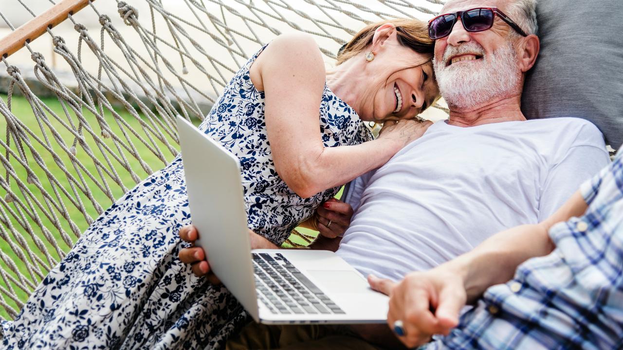 An elderly couple relaxing on a hammock and smiling with a laptop.
