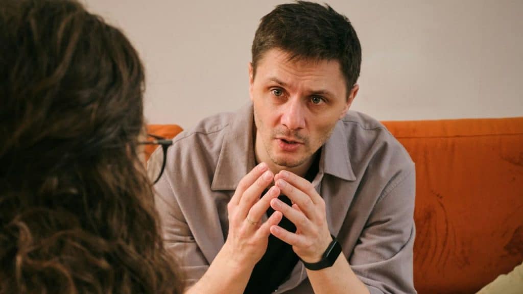 A man in a grey jacket gestures with his hands while speaking to another person.