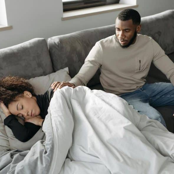 A man sitting on a couch gently touching a woman who is sleeping under a blanket.