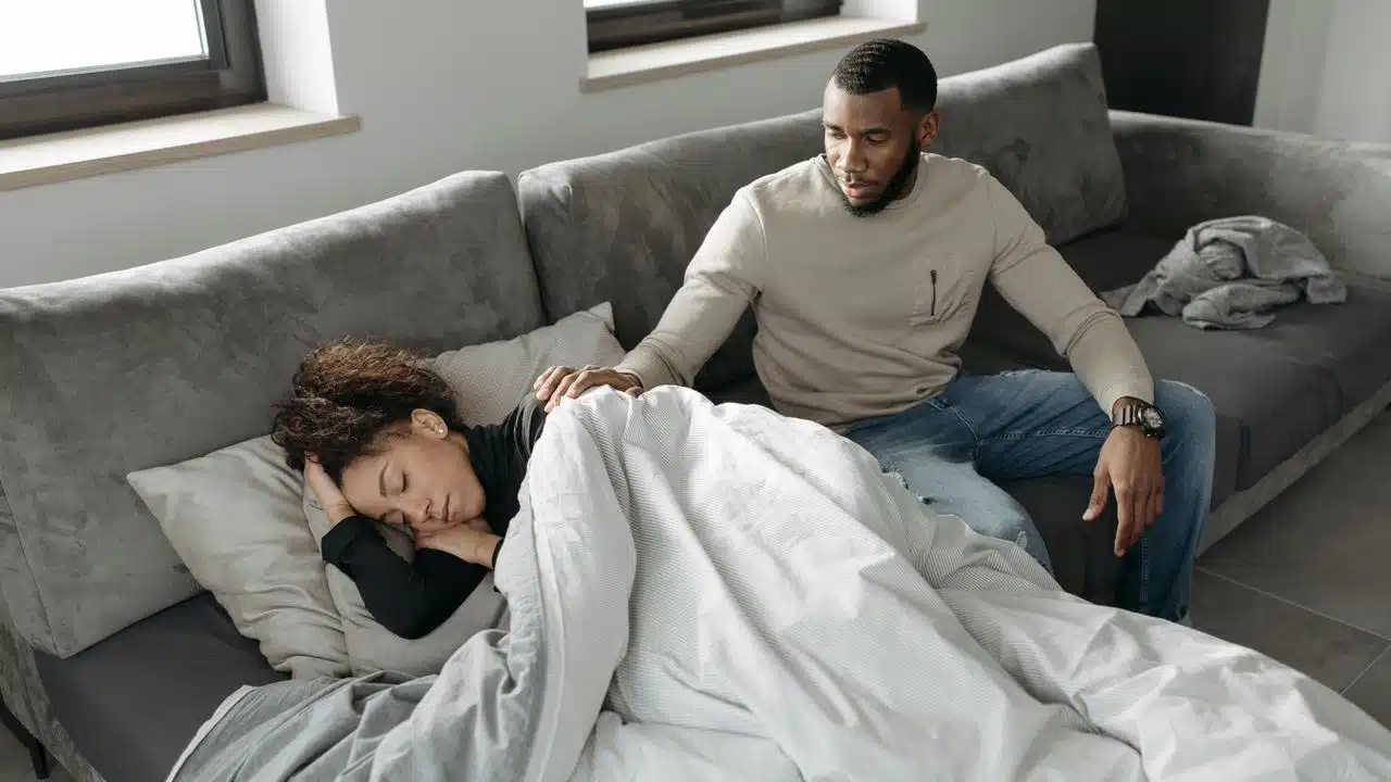 A man sitting on a couch gently touching a woman who is sleeping under a blanket.