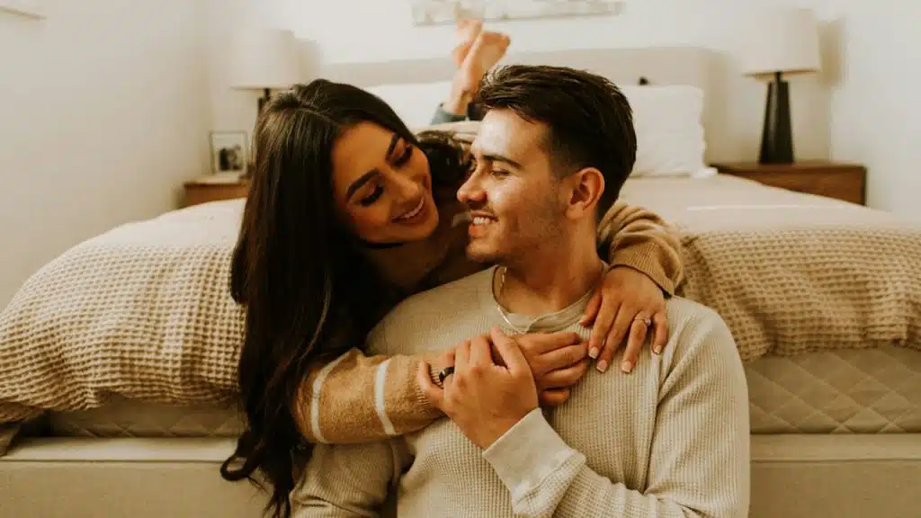A woman lying on the bed is hugging her husband from behind, who is sitting on the floor.